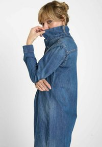 Robe chemise en denim bleu foncé avec un col classique, des manches longues et des fentes latérales. Le tissu présente une texture décolorée.