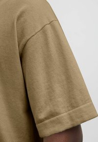 Kortærmet beige t-shirt med en blød tekstur, rund halsudskæring og rullekantdetalje, der delvist viser skulderområdet.