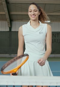 Vestuário de ténis branco com colarinho e logótipo, saia evasé, e tecido texturado; jogadora segurando uma raquete laranja perto de uma rede num court.