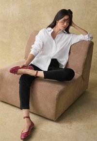 Ballerines en cuir rouge avec des pois blancs, avec une bride à la cheville, portées avec un pantalon noir et une chemise blanche à boutons, assises sur un fauteuil texturé beige.