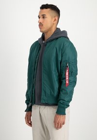 Groene bomberjack van glanzende stof met ribgebreide manchetten, ritszakken en een rode labelaccent op de mouw. Draag het over een grijze hoodie.