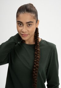 Lang, bruin gevlochten haar, draagt een donkergroene shirt met lange mouwen en een ronde halslijn, tegen een eenvoudige witte achtergrond.