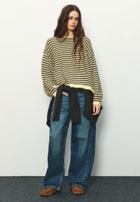Žltý a tmavohnedý pruhovaný veľký sveter v kombinácii so širokými modrými džínsami. Tmavý sveter zviazaný okolo pása, hnedé topánky.