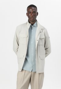 Light beige jacket with a zip closure, four front pockets, and long sleeves. Worn over a light blue button-up shirt and beige trousers.