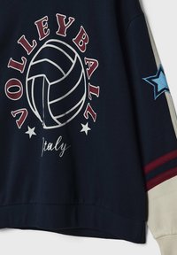Sweat-shirt bleu marine avec graphique de volley-ball, texte "VOLLEYBALL Italy" et manche crème avec étoile bleue et rayures rouges.