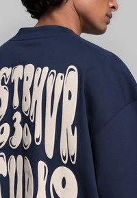 Marineblauwe sweater met een losse fit, voorzien van grote, creme-kleurige tekst op de achterkant. De stof lijkt zacht met een gladde textuur.