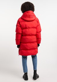 Cappotto puffer rosso con cappuccio, design trapuntato e isolamento visibile. Presenta la scritta "FRIEDEN" sul cappuccio. Indossato sopra jeans e stivali neri.