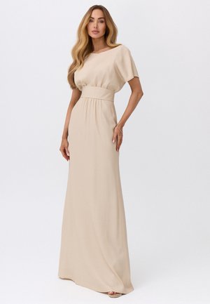 Kobieta z długimi falującymi blond włosami, ubrana w beżową suknię do podłogi z dopasowaną talią i krótkimi rękawami, stojąca na jednolitym tle.