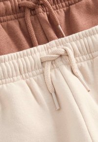 Elastic waistbands with drawstrings on beige and light brown cotton sweatpants, showing close-up fabric texture and stitching details.