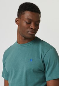 Groene katoenen t-shirt met gespikkelde textuur. Heeft een blauw geborduurd boomdetail op de borst en een klassieke ronde hals.
