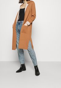 Larga chaqueta marrón con un cuello marrón claro, aberturas laterales y bolsillos de parche, combinada con unos jeans azules y botas negras de tobillo.