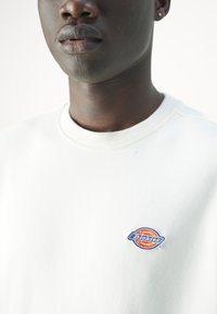 Sudadera blanca de algodón con cuello acanalado. Presenta un pequeño logo bordado de "Dickies" en colores multicolores en el pecho. Textura suave.