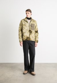 Fleecjas met een textuur in olijfgroen, met ritssluiting en borstzak. Gepaarde met zwarte broeken en taupe schoenen.