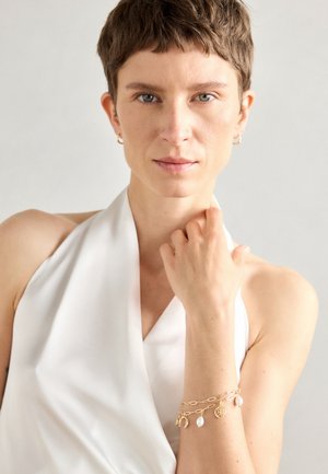 Pulseira de ouro com um design de corrente ligada e pendentes, incluindo uma concha texturizada e um pendente circular, usada em um braço nu.