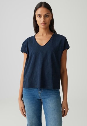 Vrouw draagt een donkerblauwe V-hals top met korte mouwen en lichtblauwe jeans, staand tegen een effen lichte achtergrond.