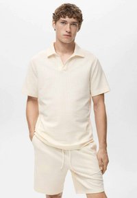 Polo shirt corto a maniche corte di un beige chiaro con tessuto testurizzato e colletto. Abbinato a pantaloni corti coordinati per un look armonioso.