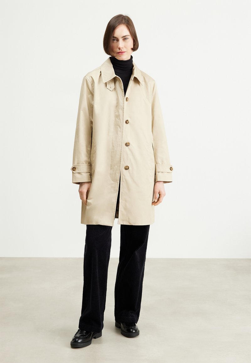 Trench de agua beige con collar clásico, cierre de botones y bolsillos laterales, combinado con pantalones negros de canalé y zapatos.