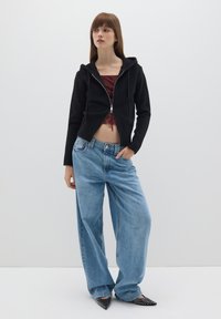 Felpa nera con zip e polsini a costine, indossata sopra una maglietta bordeaux, abbinata a jeans azzurri larghi e scarpe nere a punta con borchie.