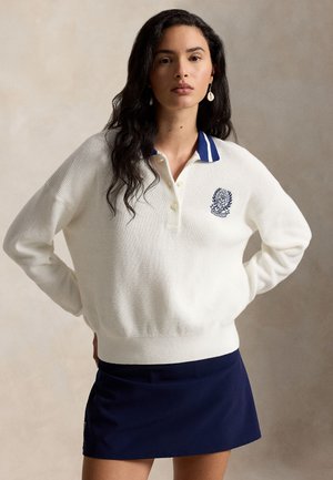 Femme aux cheveux longs et foncés portant un pull en maille blanc avec un col bleu et un blason, ainsi qu'une jupe bleu marine, debout les mains sur les hanches.