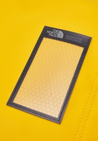 Yellow backpack material with a textured surface, featuring a black rectangular pocket with a honeycomb pattern and brand logo at the top.