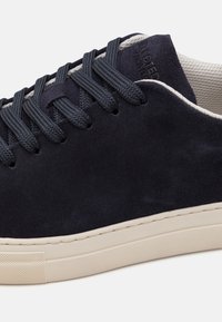 Adidași din suede bleu marine, cu șireturi plate de culoare închisă, talpă din cauciuc crem și căptușeală texturată. Detaliul cusăturii de-a lungul tălpii adaugă un contrast subtil.