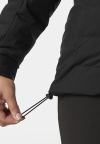 Czarna izolowana kurtka z ściągaczem u dołu. Teksturowany materiał ma matowe wykończenie. Ręka dostosowuje zamek ściągacza.