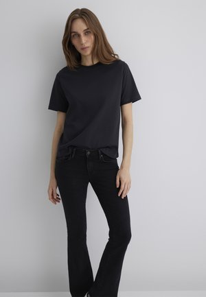 Woman standing against a gray wall wearing a black short-sleeve t-shirt and black flared jeans, looking at the camera with a neutral expression.