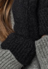 Sciarpa nera lavorata a maglia con un motivo spesso e testurizzato, posizionata su un maglione grigio a costine. Materiali morbidi e caldi sono in evidenza.