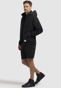 Chaqueta negra impermeable con capucha, cierre frontal y bolsillos laterales, combinada con pantalones cortos negros y zapatillas altas.