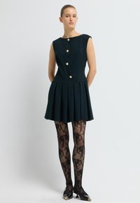 Robe sans manches bleu marine avec une jupe évasée, quatre boutons dorés, associée à des collants transparent noirs à motif floral et des chaussures noires pointues.