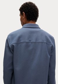 Modrá strukturovaná bunda s límcem, s viditelnými švy a uvolněným střihem. Pohled z zádu zdůrazňuje detail šití a vzor látky.