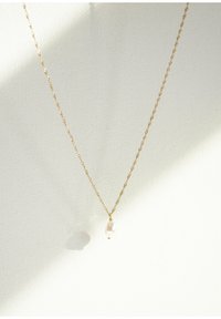 Collar de cadena trenzada dorada con un solo colgante en forma de gota de perla blanca, reposando sobre un fondo texturizado claro.