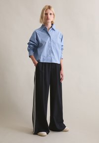 Lichtblauw cropped shirt met knoopsluiting aan de voorkant en een enkel borstzakje, gecombineerd met zwarte broek met wijde pijpen en witte strepen aan de zijkant.