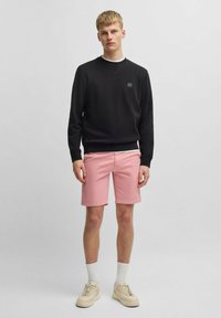 Sudadera negra con un pequeño logo, pantalones cortos rosas, calcetines blancos hasta el tobillo y zapatillas beige. Textura suave, diseño casual, pantalones cortos hasta la rodilla.