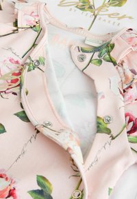 Tutina rosa con motivo floreale per neonati con bottoni a pressione parzialmente aperti, che mostrano l'etichetta interna e un motivo stampato "Baker by Ted Baker".