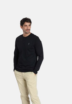 Chemise henley noire à manches longues avec trois boutons au col et un petit logo sur la poitrine, associée à un pantalon chino de couleur claire.