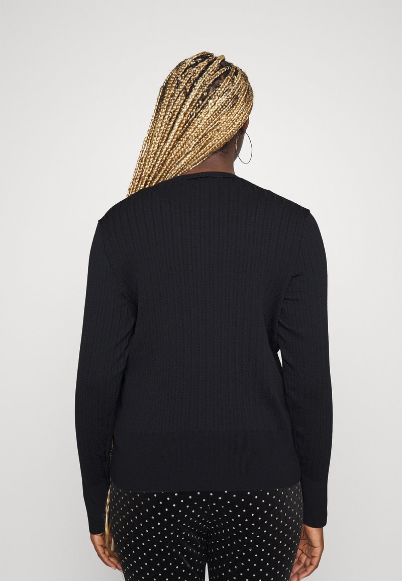 Femme aux longues tresses blondes portant un pull côtelé noir et un pantalon noir à pois blancs, vue de dos sur un fond uni.