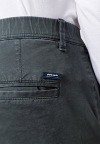 Tmavě šedé kalhoty s hladkou strukturou a zadní kapsou. Obsahují modrou etiketu s bílým textem. Úzký střih s prošitým pasem.