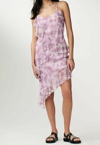 Frau in einem ärmellosen pinken Batik-Rüschenkleid mit asymmetrischem Schnitt und schwarzen, mit Nieten besetzten Sandalen, die vor einem hellen Hintergrund steht.