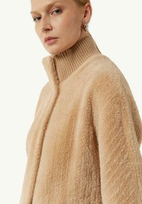 Cardigan fuzzy color tan con colletto alto a coste, caratterizzato da una texture morbida e design aperto, adatto per essere indossato a strati nelle condizioni più fresche.