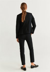 Blazer noir sur mesure avec une silhouette ajustée et un tissu légèrement texturé, associé à un pantalon noir slim et des mocassins.