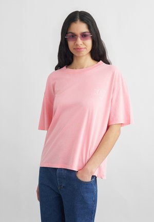 Kobieta z długimi ciemnymi włosami, nosząca różowe okulary przeciwsłoneczne, różowy oversize'owy t-shirt i niebieskie dżinsy, stojąca z jedną ręką w kieszeni.