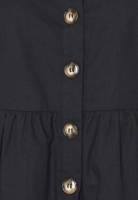 Svart klänning med knäppt framtill och fyra stora, rundade knappar. Tyget har en slät textur med en samlad fålldetalj.