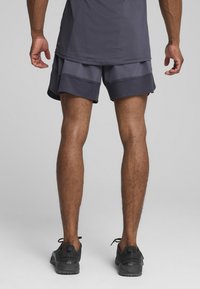 Grå idrottsshorts med lagerdesign, med en strukturerad midja. Bärs med svarta träningsskor, sedd från baksidan.