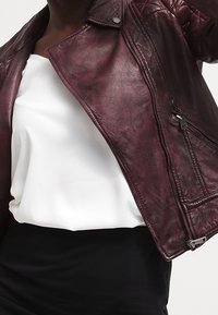 Giacca in pelle bordeaux con dettagli trapuntati sulle spalle, zip in argento e design aderente, indossata sopra una maglietta bianca e una gonna nera.