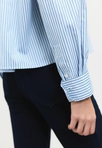 Gestreept blauw overhemd met lange mouwen, voorzien van witte en marineblauwe strepen, een gebogen zoom en manchetdetail. Gepaard met donkere broeken.