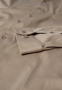 Beige skjorte med knapper og en glat tekstur, der har brune knapper og opfoldede manchetter. Stoffet er let krøllet, hvilket fremhæver dets blødhed.