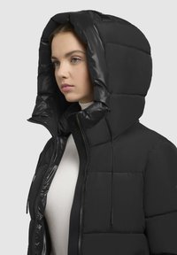 Giacca a vento nera con un grande cappuccio imbottito. Presenta una finitura lucida, chiusura con zip e texture trapuntata. Dettagliata con lacci.