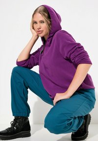 Sudadera con capucha morada y cremallera, combinada con pantalones jogger en color teal y botas negras de tobillo. El conjunto presenta un diseño informal y relajado.