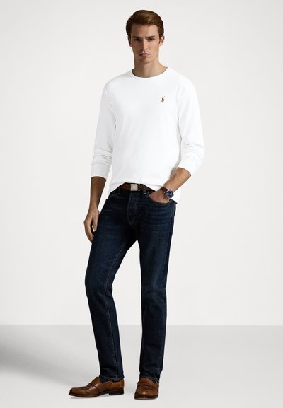 Fehér hosszú ujjú póló logóval, sötétkék farmer, barna elegáns cipő és egy óra. Egyszerű design, testhezálló szabás, sima textúra.